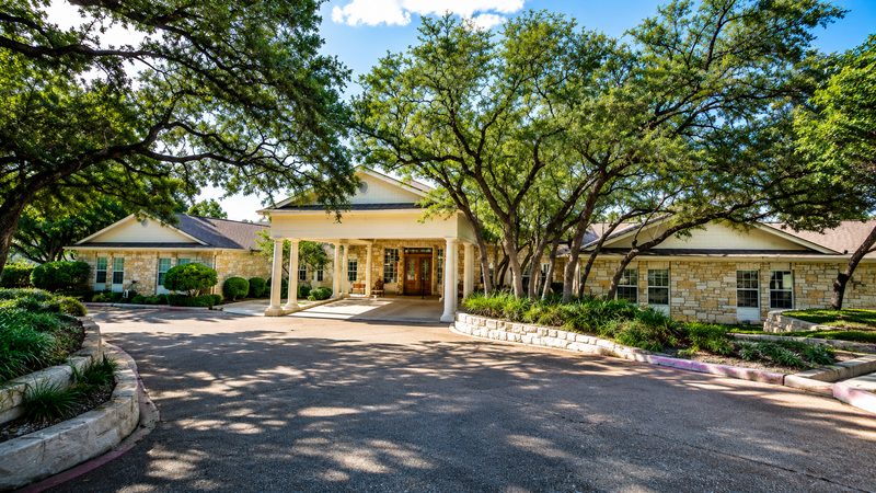 Front entrance at Bee Cave, a limestone portico with white columns under live oaks, circular drive in front