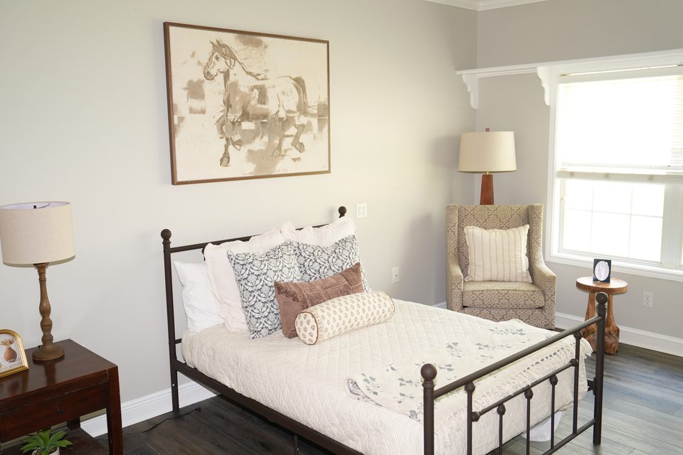 Private suite at Bee Cave with a made bed, a framed print, a wing chair, and a window onto the trees